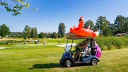 Flamingo-uimarengas golfauton katolla, autossa äiti ja lapsia. Golfbil med flamingoformad badring på taket och mamma med barn