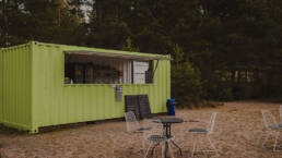 Vihreä kahvilakontti rannalla, edustalla pöytiä ja tuoleja. En grön cafécontainer på stranden, framför den står stolar och bord.